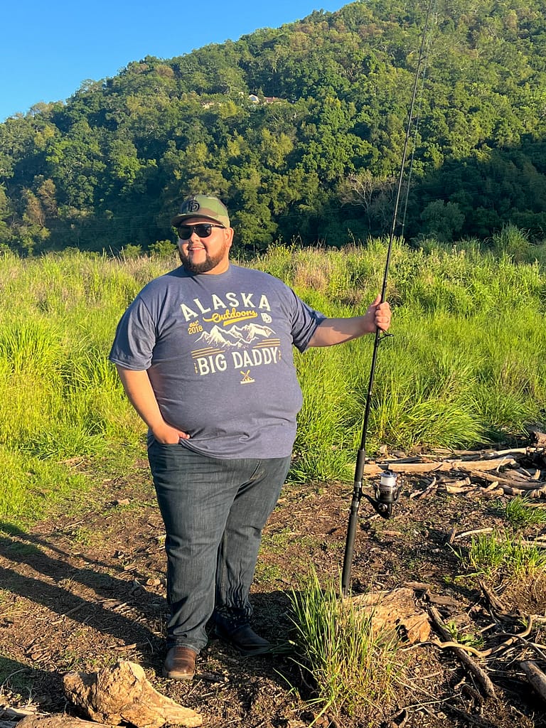 A man holding a fishing pole wearing the Big Daddy Alaska Outdoors Tee in Heather Navy in size 6XL and the Big Daddy Puff Logo Snapback hat in Camo Color